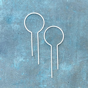 May include: Two white, minimalist earrings with a circular top and a straight, thin post. The earrings are set against a textured, teal-colored background, creating a clean and modern aesthetic. These earrings are a simple yet elegant accessory.