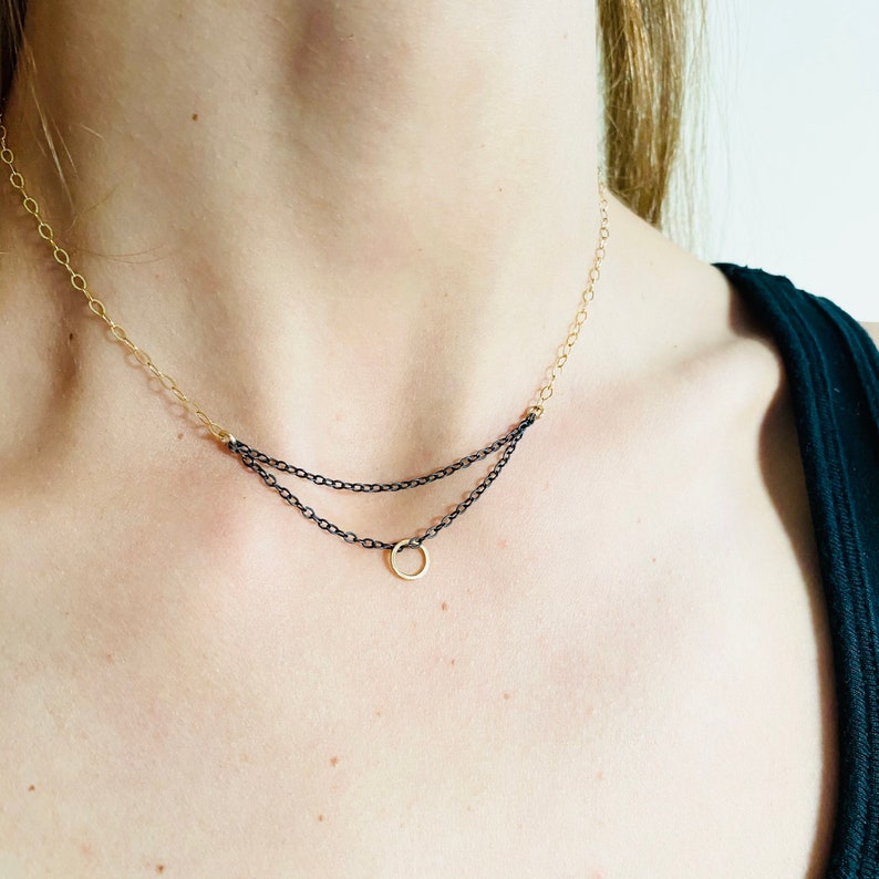 May include: A layered necklace with a gold-tone chain and a black chain. The necklace features a small gold circle pendant. The necklace is worn on a person.