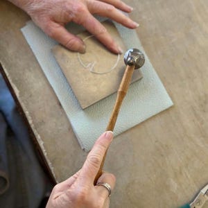 May include: A jewelry-making scene featuring a small metal hammer with a wooden handle. The hammer is poised above a metal piece on a light-colored surface. A hand is holding the hammer, and another hand is working on the metal.