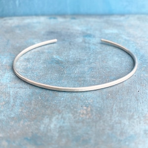 May include: A simple, silver-colored cuff bracelet. The open-ended bangle is round and thin, with a smooth, matte finish. The jewelry is displayed against a light blue, textured background.