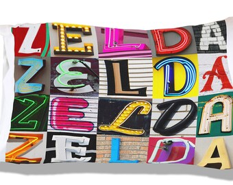 Op de afbeelding: Een wit kussen met een kleurrijke collage van de letters "ZELDA" gemaakt van verschillende neon- en vintage borden. De letters zijn in verschillende kleuren, lettertypen en stijlen, wat een levendig en eclectisch ontwerp creëert.