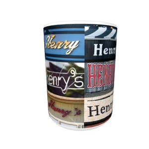 Op de afbeelding: Witte keramische mok met een collage van kleurrijke borden met de naam "Henry's" in verschillende lettertypen en stijlen.