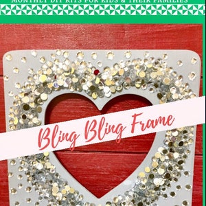 May include: A white wooden frame with a heart-shaped cutout, decorated with silver and gold glitter. The frame is on a red wooden background. The text "Bling Bling Frame" is written in pink on a white banner.