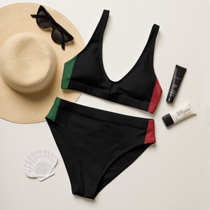 May include: Black bikini with green and red trim. The top is a V-neck design, and the bottoms are high-waisted. A sun hat, sunglasses, sunscreen, and a seashell are also visible in the image.