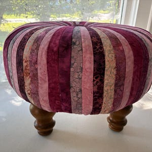 May include: A round ottoman featuring a tufted top and vertical fabric panels in various shades of pink, magenta, and deep purple. The fabric displays floral and marbled patterns. The piece is supported by four short, turned wooden legs in a brown finish.