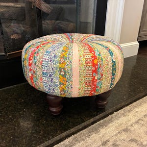 May include: A round ottoman featuring a patchwork design of vertical fabric strips in various floral and geometric patterns, including shades of red, blue, yellow, green, and pink. The piece is supported by four dark brown, turned wooden feet.