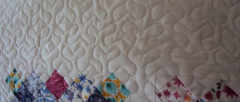 May include: White quilted fabric with a wavy pattern. The fabric is bordered with colorful patchwork squares in shades of pink, blue, green, and yellow.