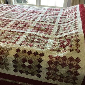 Puede incluir: Un edred&oacute;n hecho a mano con un fondo crema y un patr&oacute;n repetitivo de cuadrados rojos y marrones. El edred&oacute;n tiene un borde rojo ancho y est&aacute; sobre una cama. El dise&ntilde;o del edred&oacute;n es de estilo patchwork tradicional.