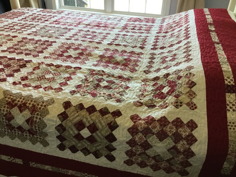 Puede incluir: Un edred&oacute;n hecho a mano con un fondo crema y un patr&oacute;n repetitivo de cuadrados rojos y marrones. El edred&oacute;n tiene un borde rojo ancho y est&aacute; sobre una cama. El dise&ntilde;o del edred&oacute;n es de estilo patchwork tradicional.