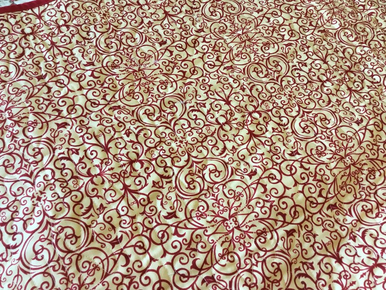 Puede incluir: Tela color crema con un patr&oacute;n repetitivo de volutas y dise&ntilde;os florales burdeos. El patr&oacute;n se distribuye uniformemente por la superficie, creando una textura visualmente rica y detallada. La tela parece estar acolchada.