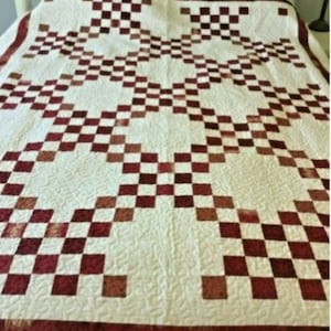 May include: A handmade quilt with a cream-colored background and a repeating pattern of burgundy and brown squares. The quilt features a border of burgundy fabric and is likely made of cotton or a similar material. The design is a classic, geometric pattern.