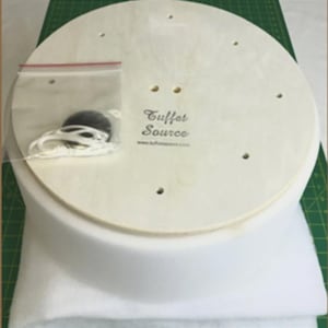May include: A round, light-colored wooden base with pre-drilled holes, labeled "Tuffet Source." A small plastic bag contains black buttons and white string. The base sits atop a white, rounded cushion.