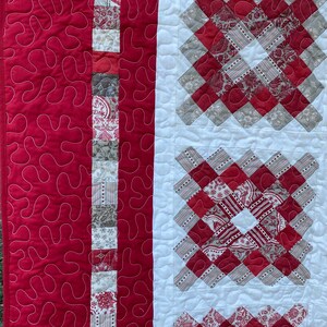 Puede incluir: Primer plano de un edred&oacute;n hecho a mano con un dise&ntilde;o de patchwork. El edred&oacute;n presenta una tela roja vibrante con patrones de remolino blancos y una secci&oacute;n central con cuadrados estampados rojos y grises. Una tira vertical de tela estampada a&ntilde;ade detalles.