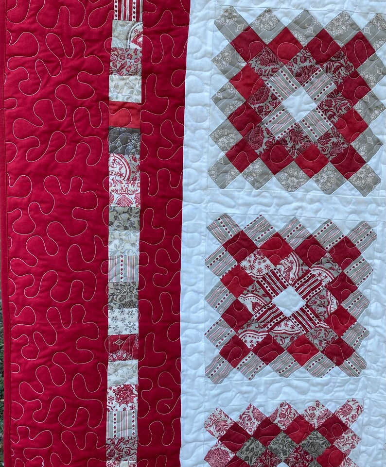 Puede incluir: Primer plano de un edred&oacute;n hecho a mano con un dise&ntilde;o de patchwork. El edred&oacute;n presenta una tela roja vibrante con patrones de remolino blancos y una secci&oacute;n central con cuadrados estampados rojos y grises. Una tira vertical de tela estampada a&ntilde;ade detalles.