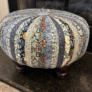 May include: A round ottoman featuring vertical fabric panels with various floral and botanical patterns in shades of blue, cream, orange, and green. The top center has a fabric-covered button, and the base is supported by four dark brown wooden feet.