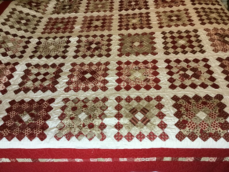 Puede incluir: Un edred&oacute;n hecho a mano con un patr&oacute;n repetido de bloques cuadrados en tonos de rojo, crema y beige. Los bloques est&aacute;n dispuestos en una cuadr&iacute;cula, con un borde rojo en la parte inferior. El edred&oacute;n probablemente est&aacute; hecho de algod&oacute;n.