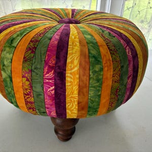 May include: A round ottoman featuring a tufted center button and vertical fabric panels in alternating shades of purple, orange, and green with floral patterns. The upholstered seat is supported by a single dark brown turned wooden leg.