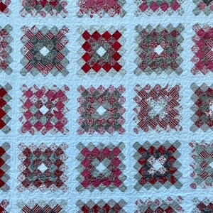 Puede incluir: Primer plano de un textil acolchado con un patr&oacute;n repetitivo de bloques cuadrados. Cada bloque est&aacute; compuesto por cuadrados m&aacute;s peque&ntilde;os en tonos de rojo, gris y blanco, creando un dise&ntilde;o geom&eacute;trico. El edred&oacute;n tiene un fondo blanco.