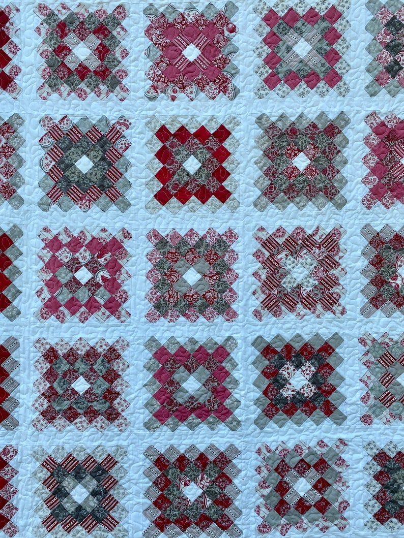 Puede incluir: Primer plano de un textil acolchado con un patr&oacute;n repetitivo de bloques cuadrados. Cada bloque est&aacute; compuesto por cuadrados m&aacute;s peque&ntilde;os en tonos de rojo, gris y blanco, creando un dise&ntilde;o geom&eacute;trico. El edred&oacute;n tiene un fondo blanco.