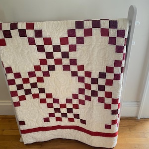 May include: A handmade quilt with a checkerboard pattern in shades of red and burgundy on a cream background. The quilt is displayed on a white wooden rack, showcasing its intricate design and craftsmanship. The quilt's border is a solid red stripe.