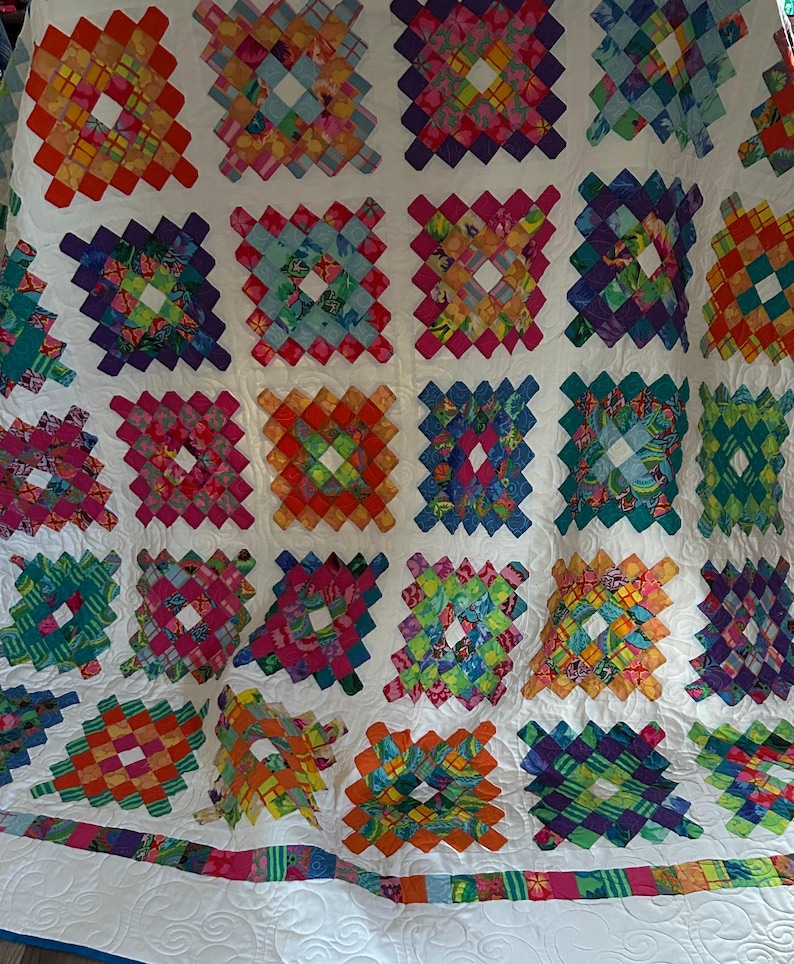 May include: A colorful quilt with a white background and a diamond pattern. The quilt is made up of many different fabrics, each with a unique design. The quilt is bordered with a white trim and a colorful ribbon.