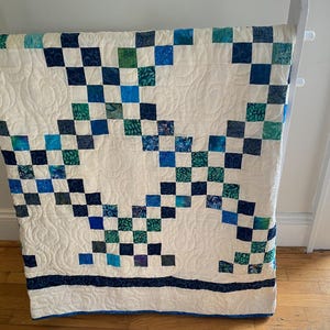 Puede incluir: Un edredón hecho a mano con un patrón de tablero de ajedrez en tonos de azul y verde sobre un fondo crema. El edredón está colgado en un estante blanco, mostrando su intrincado diseño y artesanía. Las dimensiones del edredón no son visibles.