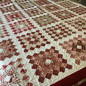 Puede incluir: Un edred&oacute;n hecho a mano con un patr&oacute;n repetitivo de formas de diamante rojas y blancas. El edred&oacute;n presenta varios tonos de telas rojas y blancas, creando un dise&ntilde;o texturizado y visualmente atractivo. Un art&iacute;culo de cama acogedor y decorativo.