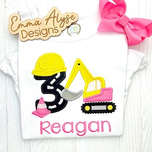 May include: A white t-shirt featuring an embroidered design of a black number three shaped like a road, topped with a yellow hard hat, a yellow and pink excavator, and a pink traffic cone. The name Reagan is embroidered in pink below the design.