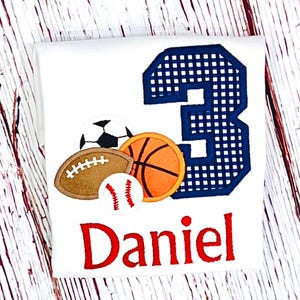 May include: A white t-shirt featuring an embroidered design with a large navy blue plaid number three, a soccer ball, a brown football, an orange basketball, and a white baseball with red stitching. The name Daniel is embroidered in red below.