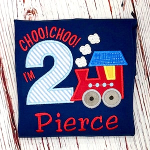 May include: Navy blue toddler shirt with embroidered text and train graphic. The shirt says "CHOO! CHOO! I'M 2 Pierce" in red and light blue. The train is red, blue, yellow, and gray.