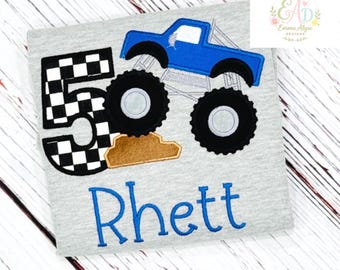 May include: A heather grey t-shirt featuring an embroidered applique of a blue monster truck driving over a brown dirt mound. To the left is a large number five with a black and white chequered pattern. The name Rhett is embroidered in blue below.