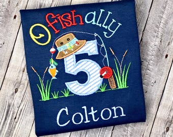 May include: A navy blue t-shirt featuring an embroidered design with the text "Fishally 5" and "Colton". The graphic includes a brown hat, a blue and white striped number five, a fishing rod, a small orange fish, and green grass blades.
