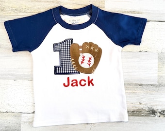 May include: White baseball-themed t-shirt with navy blue sleeves. The shirt features a baseball glove and ball design, a gingham number 1, and the name "Jack" in red lettering. A fun and sporty design for a young child.