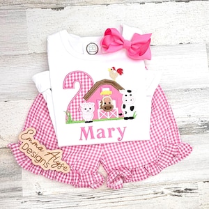 May include: A white two-piece outfit with a pink gingham pattern. The top features a farm scene with a number 2, a barn, and farm animals. The name "Mary" is embroidered below the design. A large pink bow and wooden sign are included.