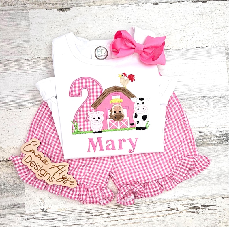 May include: A white two-piece outfit with a pink gingham pattern. The top features a farm scene with a number 2, a barn, and farm animals. The name "Mary" is embroidered below the design. A large pink bow and wooden sign are included.