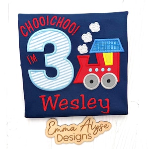 May include: A navy blue t-shirt featuring an embroidered design of a red train with a blue roof, white smoke clouds, and the text "CHOOI CHOO! I'M 3" in red and light blue stripes. The name "Wesley" is embroidered in red below the train graphic.