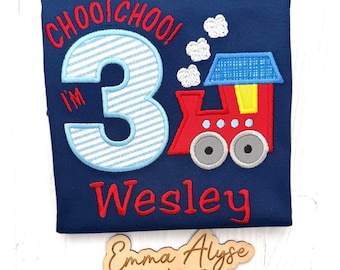 May include: A navy blue t-shirt featuring an embroidered design of a red train with a blue roof, white smoke clouds, and the text "CHOOI CHOO! I'M 3" in red and light blue stripes. The name "Wesley" is embroidered in red below the train graphic.