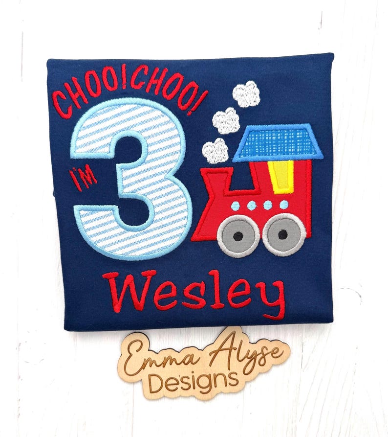 May include: A navy blue t-shirt featuring an embroidered design of a red train with a blue roof, white smoke clouds, and the text "CHOOI CHOO! I'M 3" in red and light blue stripes. The name "Wesley" is embroidered in red below the train graphic.