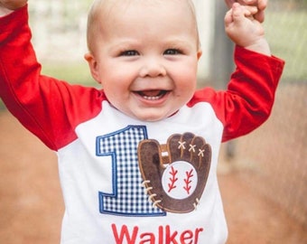May include: A white and red baseball-themed shirt with the number "1" in blue gingham and a brown baseball glove applique. The name "Walker" is embroidered in red. The child is smiling and being held up by an adult's hands.