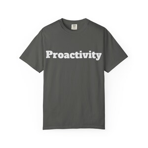 May include: A dark gray t-shirt with the word "Proactivity" printed in bold, white letters across the front. The shirt has short sleeves and a crew neck. The fabric appears to be a soft cotton blend.