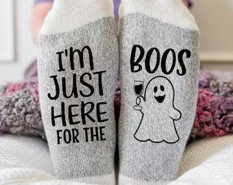 May include: A pair of grey and white socks featuring black text and a graphic. The left sock reads I'M JUST HERE FOR THE and the right sock features the word BOOS above a cartoon ghost holding a wine glass. The socks have white toe and heel sections.