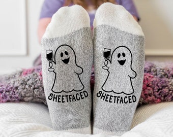 May include: A pair of heather gray socks with white cuffs, featuring a black graphic of a ghost holding a wine glass and the text "SHEETFACED" printed in a bold, sans-serif font on the front of each sock, positioned side by side.