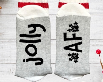 May include: A pair of light gray crew socks with white and red striped cuffs. The left sock features the black text "jolly" in a script font, while the right sock displays the black text "AF" flanked by black holly leaf icons.