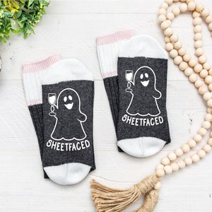 May include: A pair of dark gray socks with white heels and toes, featuring a white graphic of a ghost holding a wine glass and the text "SHEETFACED" printed in white. The socks have a ribbed cuff with a light pink stripe at the top edge.