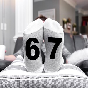 May include: A person is relaxing on a couch with their feet up, wearing gray socks with the numbers 6 and 7 printed on them. A white mug sits nearby. The socks are the focal point, with a neutral background.