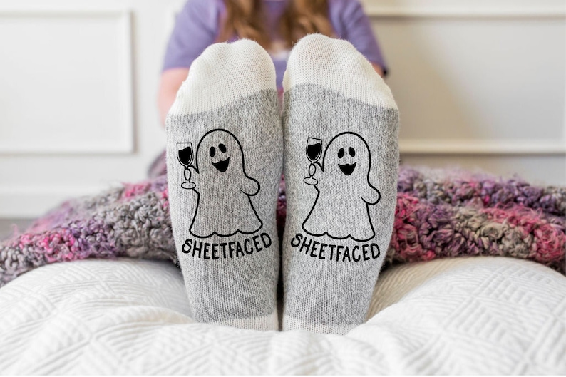 May include: A pair of heather gray socks with white cuffs, featuring a black graphic of a ghost holding a wine glass and the text "SHEETFACED" printed in a bold, sans-serif font on the front of each sock, positioned side by side.