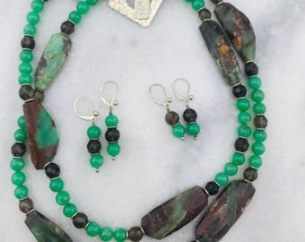 Op de afbeelding: Een ketting- en oorbellenset met groene, bruine en zwarte kralen. De ketting heeft grote, onregelmatig gevormde kralen en een zilveren sluiting. De oorbellen hebben een vergelijkbaar ontwerp met kleinere kralen en zilveren haken.