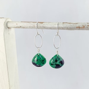 May include: A pair of silver dangle earrings with green and black gemstone drops. The earrings feature a simple oval hoop at the top.