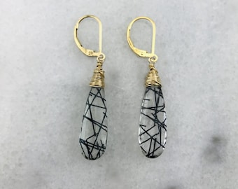 May include: A pair of gold earrings with teardrop-shaped black tourmaline stones. The stones are clear with black inclusions that resemble lines or veins.