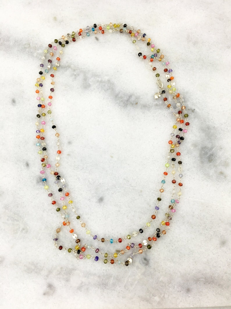 May include: A multi-strand necklace with small, colorful gemstone beads. The necklace is made of silver chain and features a variety of colors, including red, orange, yellow, green, blue, purple, and pink.